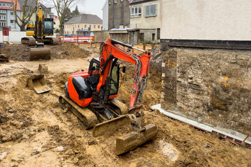 A small red excavator at a construction site is digging a pit in the dirty ground.