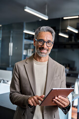 Happy mature older business man executive standing in office using digital tablet. Middle aged...