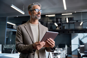 Middle aged business man professional executive standing in office using tablet looking away. Busy...
