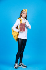 Adorable teen girl in polka dot bow demonstrating bunch of books, tied with twine, wistfully looking at camera. Front view of cute female student showing books, isolated on blue. Concept of studying. 