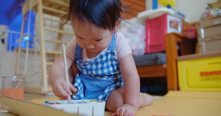 An engrossed toddler in a blue checkered dress discovers creativity with a paintbrush in hand and...