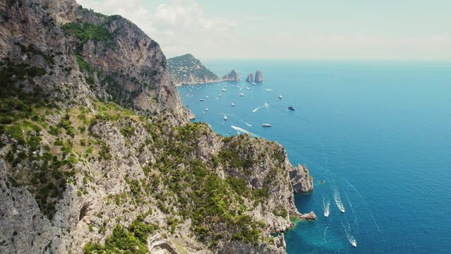 Lush green cliffs dive into a sapphire sea dotted with boats. Seascape with towering cliffs Faraglioni. Capri Island, Italy.