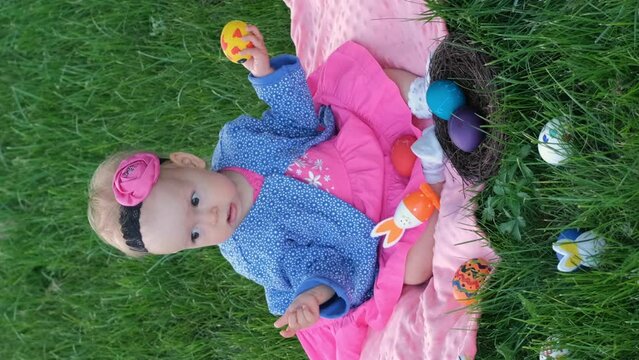 Vertical video. Cute baby girl playing with colorful Easter eggs on grass