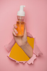 A woman's hand shows a bottle of cleansing foam through a hole in a paper background. - 738247382