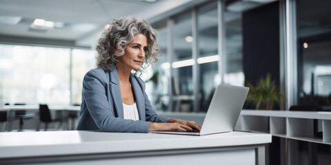 Elegant senior lady in blue blazer typing on laptop at modern workplace. Corporate style concept. Generative AI - obrazy, fototapety, plakaty