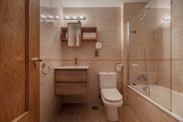 Front image of a classic bathroom, with a marble-style tile wall with a border, a small hanging wooden cabinet with an integrated mirror and a bathtub with a folding glass screen - obrazy, fototapety, plakaty