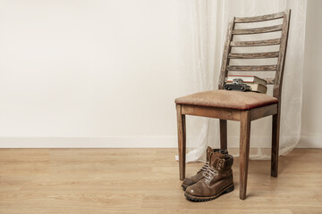 Brown leather hiking boots resting under a emaciated wooden chair with an old camera resting on the back along with some books