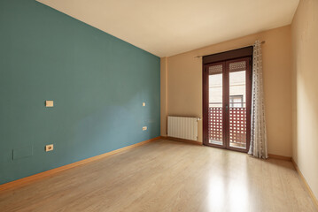 An empty room with a blue painted wall, a balcony with double glass and brown aluminum doors with a curtain, a white aluminum radiator and light wooden flooring
