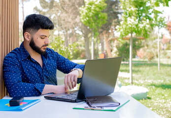 Front view of handsome asian freelancer sitting at table, working outdoors. Brunette young male with beard using laptop, looking at watch. Concept of youth and urban life.