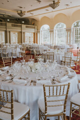 
Salón de celebraciones repleto de mesas decoradas en tonos blancos y sillas doradas. Mesas redondas con manteles blancos, cubertería y vasos de cristal transparente.