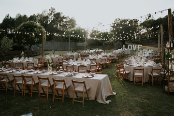 
Fotografía muy general de un banquete de bodas al aire libre. Mesas diferentes, alargadas y redondas decoradas. Entorno natural, árboles y césped, iluminado con guirnaldas. En un extremo un cartel.