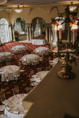 
Plano general. Salón de celebraciones repleto de mesas decoradas en tonos blancos y sillas doradas. Alfombra granate y lámpara de araña en primer plano.