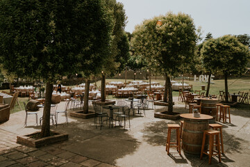 
Fotografía muy general de un banquete de bodas al aire libre. Mesas diferentes, alargadas y redondas decoradas. Entorno natural, árboles y césped, iluminado con guirnaldas. 