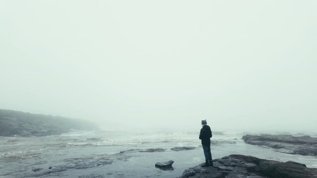 Young boy standing on the rocks on a coastal landscape. Winter scene with mist and fog. Seascape, outdoor clothing, crashing waves and rough seas