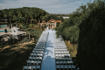 
Plano general de una ceremonia judía antes de su comienzo. Vemos multitud de sillas de madera blanca, colocadas en dos filas, separadas por una alfombra blanca. Encima de las sillas hay una kipá.