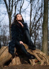 Beautiful young woman in fur coat sits on log in the forest