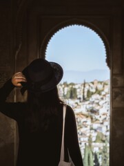 A beautiful young girl in a dress and a hat looking at the Albaicin district of Granada from the window of the Alhambra palace