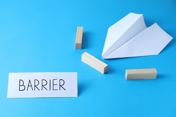 Paper plane movement blocked by wooden blocks and card with word Barrier on light blue background....