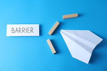 Paper plane movement blocked by wooden blocks and card with word Barrier on light blue background, flat lay. Development through obstacles overcoming - obrazy, fototapety, plakaty