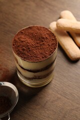 Delicious tiramisu in glass, biscuits and sieve with cocoa powder on wooden table