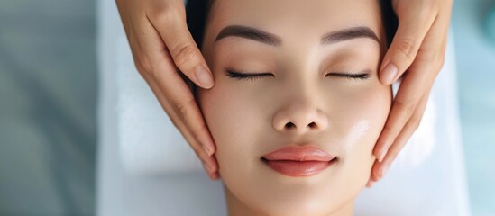 Asian woman getting facial massage with eyes closed in beauty salon. Close-up of beautician's hands.