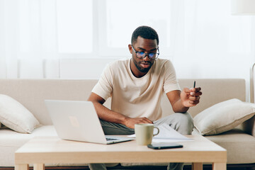 Young African American freelancer working on laptop while sitting on a modern sofa in his home He looks happy and relaxed, enjoying the comfort of his living room The room is filled with a cozy
