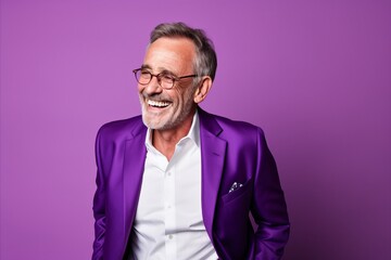 Portrait of a happy senior man in a purple suit and glasses smiling on a purple background.