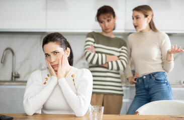 Offended middle-aged woman sitting in the kitchen while two women talking angrily to her