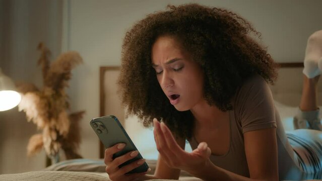 Upset shocked sad African American girl lady disappointed ethnic woman worried phone crash mobile error mistake scam message frustrated female on bed evening home with smartphone loss failure bad news