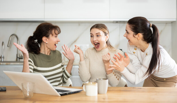 Female friends are sitting at table, reading letter on computer about invitation to study and scholarship, rejoicing at good news, are glad over victory.