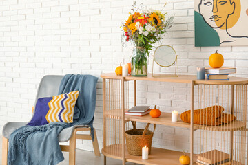 Shelving unit with bouquet of autumn flowers and armchair near white brick wall