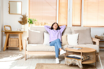Beautiful young Asian woman in stylish hoodie sitting on sofa at home