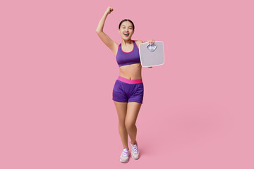 Happy young woman in sportswear and with weight scales on pink background