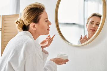 beautiful blonde woman in comfy white bathrobe applying face cream while in bathroom at home - obrazy, fototapety, plakaty