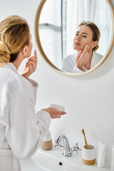 beautiful blonde woman in comfy white bathrobe applying face cream while in bathroom at home