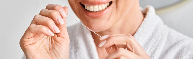 cropped view of jolly woman in bathrobe cleaning her teeth with dental floss in bathroom, banner
