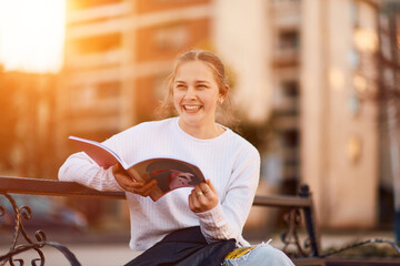 A beautiful blonde teenager sits on a bench in the park, taking a break from school to immerse herself in a book, accompanied by her school backpack