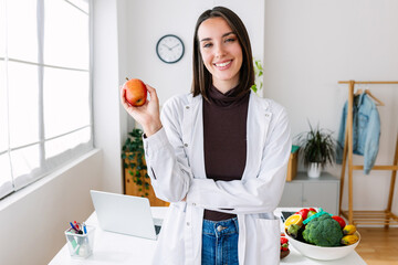 Smiling portrait of young nutritionist woman holding red organic apple at medical office. Healthy...