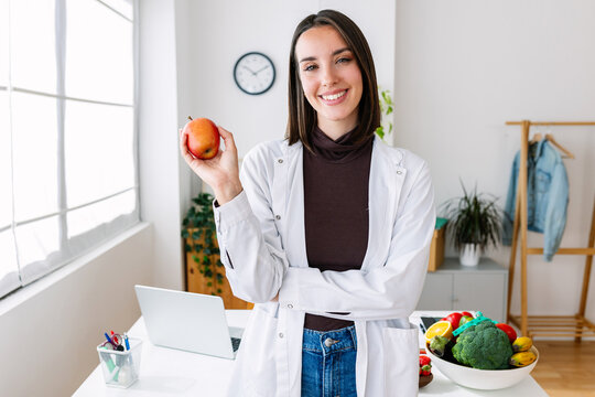 Smiling portrait of young nutritionist woman holding red organic apple at medical office. Healthy lifestyle and nutrition concept.