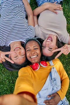Vertical selfie above of three cheerful multiracial young women lying on grass outdoor. Female looking at camera smiling and making peace sign with hands enjoying summer vacation. Generation z in park