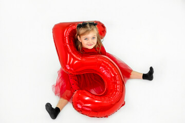 A funny girl with a big red foil balloon in the shape of the number five is sitting on the floor smiling sweetly. The child celebrates his birthday