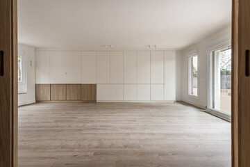 Living room of a recently renovated empty house with white painted walls and a bookcase with white wooden doors seen from the oak wooden doors