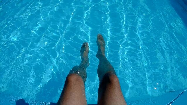 Close-up of slender male legs against the blue water by the pool on a hot summer day, under the sun. The man dangles her legs in the water of the pool, resting, sunbathing.