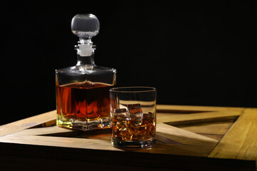 Whiskey with ice cubes in glass and bottle on wooden crate against black background, space for text