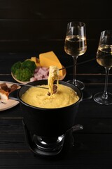 Dipping pieces of ham and bread into fondue pot with melted cheese on black wooden table