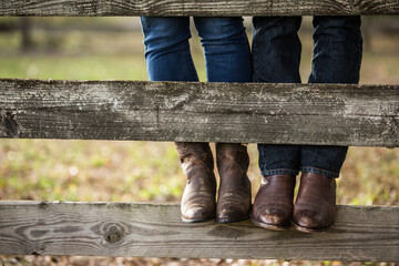 Cute Playful Close up Detail of Man and Woman model couple wearing cowboy boots and jeans standing on rustic wooden fence on farm © Betsy