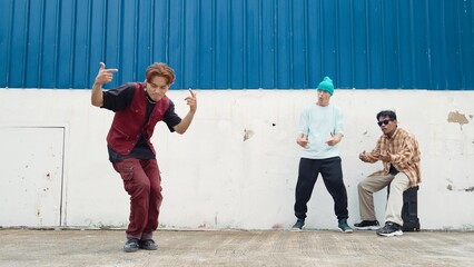 Young street dancer dancing in hip hop style with multicultural friends cheering behind at wall. Attractive handsome man stretch arms at street with blue background. Outdoor sport 2024. Endeavor.