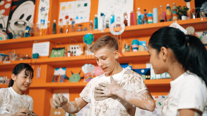 Smart caucasian teenager and happy friend with mixed races put clay on body in art lesson. Diverse...