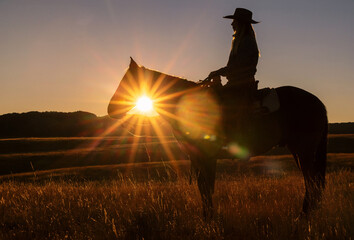 Cowgirl on a horse at sunrise making a starburst sun flare and silhouette - obrazy, fototapety, plakaty