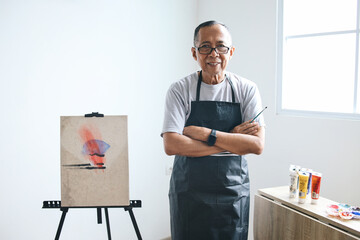 Senior Asian man wear apron smiling at camera while standing near easel at home